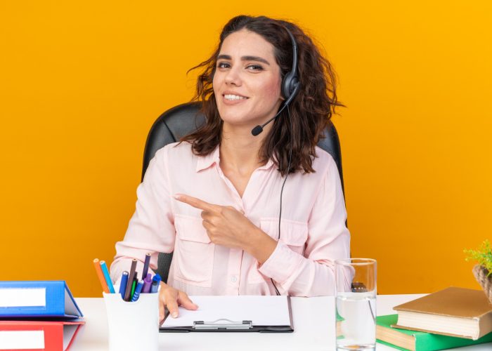 smiling-pretty-caucasian-female-call-center-operator-headphones-sitting-desk-with-office-tools-pointing-side-isolated-orange-wall